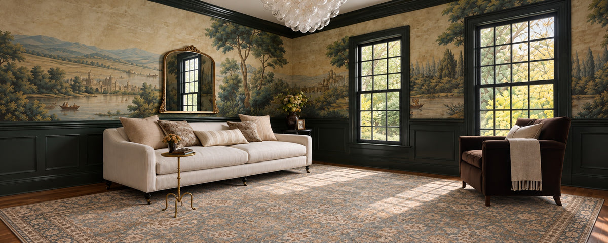 A styled interior room featuring a large cream sofa with patterned pillows, a dark brown armchair with a throw, a patterned area rug, dark green wainscoting, and extensive landscape wall murals behind the furniture.